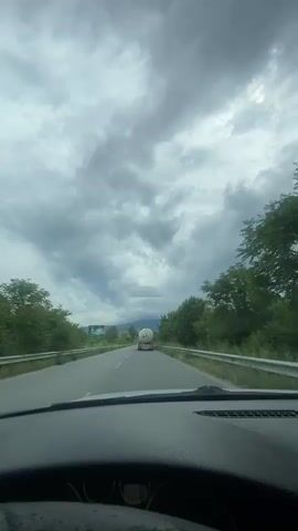 The video shows a first-person view from inside a car driving on an asphalt road. The dashboard and a portion of the steering wheel are visible in the foreground. Ahead, a white truck is seen with a large, white, spherical tank secured on its trailer. The car appears to be following this truck.

The road is lined with green trees and vegetation on both sides, with guard rails separating the road from the foliage. In the distance on the left, a billboard is partially visible. The sky is heavily overcast with dense, grey clouds, suggesting a cloudy day, possibly before or after rain. The overall scene depicts a rural or semi-rural highway. The location context provided indicates this is in Glavinitsa, Bulgaria.