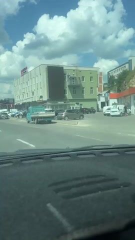 A dashcam view shows a street scene with cars and buildings. In the foreground, the textured dashboard of a vehicle is visible. The road has white lane markings. On the right side of the road, a light green building with a white facade and windows is prominent. A red sign with white lettering is on top of the building. A dark rectangular screen is attached to the building's facade. Several cars are parked along the right side of the street, and a light blue flatbed truck is driving in the street. On the left side of the street, more cars are visible, and a red truck is stopped at what appears to be an intersection. The sky is blue with fluffy white clouds.