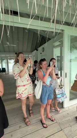 A group of women, likely tourists, stand on a wooden porch or deck under a thatched roof, all facing the same direction and holding up smartphones to seemingly take photos or videos. The woman on the left wears a black dress, the next woman is in a strawberry-patterned pajama set, the third has a blue tank top and denim shorts, and the woman on the right is in a floral dress with a beach tote. A man can be seen slightly behind them. They appear to be in a tropical location, possibly a resort or villa overlooking the ocean. The overall scene seems relaxed and vacation-like.