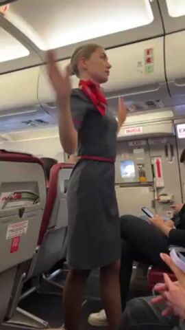 The video shows a flight attendant in a grey dress with a red belt and scarf standing in the aisle of an airplane. She is demonstrating something with her arms, then clasps her hands behind her back. Passengers are seated on either side of the aisle. The interior of the plane is visible with overhead compartments and exit doors.