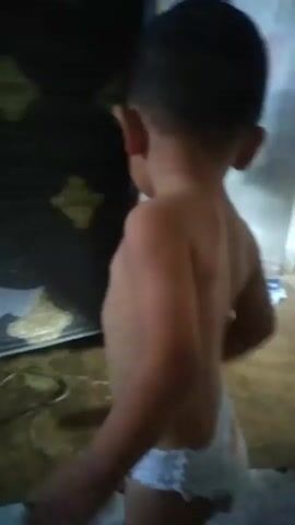This media file is a short video clip featuring a young child. The child, seen from behind, appears to be a toddler and is wearing a diaper. The setting seems to be indoors, possibly a home, with a dark doorway or panel visible on the left and a lighter wall with some vertical pink fabric hanging on the right. The floor is a neutral color, and appears somewhat wet or reflective in places. The child's back and shoulders are visible, and they have short dark hair. The lighting suggests it could be daytime, but it's difficult to ascertain specific time of day. The child is actively moving, first standing and looking away from the camera, then turning and moving slightly to the right, and finally crouching down to touch the wall. There is no discernible text or spoken language in the clip. The overall atmosphere is casual and observational, capturing a moment of a child's activity.