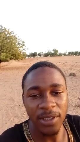 A young man with dark skin is taking a video of himself. He's wearing a black shirt with gold trim and has a short haircut. He's holding what appears to be a vape pen in his hand. The background is a sandy, desert-like landscape with sparse vegetation and a few trees in the distance. The lighting suggests it is either early morning or late afternoon. The man is smiling and seems to be singing or talking directly to the camera.