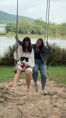 Two young women sit on a wooden swing hanging from ropes and they are swaying gently. Both are smiling and having a good time. The woman on the left wears a white dress with long balloon sleeves, a wide brown belt, and light-colored sandals. She also has a small red purse. The woman on the right wears a denim jacket, a dark top, jeans, and gray sneakers. She wears glasses. Behind them is a lake, some greenery, trees, and a building. In the distance, there is a mountain. The ground under the swing is dirt and grass.
