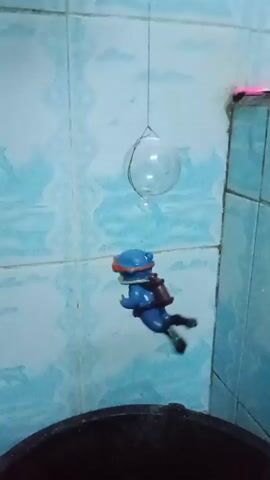 A small toy figure, dressed as a scuba diver, hangs from a string above a black bucket filled with water. The figure is initially suspended at a distance above the bucket, with a clear bubble also hanging above it. In the first few seconds, the figure begins to fall. It lands in the bucket, causing ripples on the water's surface. The clear bubble remains suspended above. The background appears to be a tiled wall.