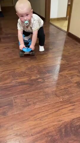 A baby with a pacifier in its mouth is kneeling on a blue skateboard on a wooden floor. The baby is wearing a light-colored shirt with a pattern and dark pants. The baby's hands are on the front of the skateboard. The baby appears to be attempting to move forward on the skateboard. The lighting suggests an indoor setting. The floor is a dark brown wood laminate. The background is out of focus but appears to be a hallway with a doorway.