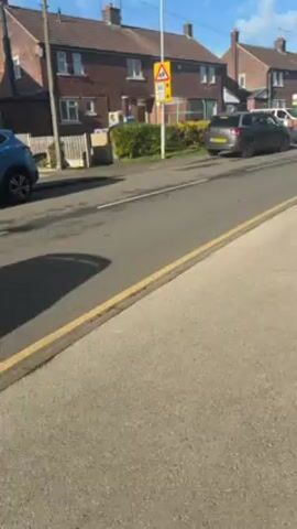 The video shows a street in a residential area with red brick houses on either side.  A 20 miles per hour school crossing sign is visible. Two parked cars are present; a gray SUV and a silver sedan, along with a white van further down the street. The road is asphalt, with a yellow curb marking the edge of the sidewalk where the video is taken. The sky is blue with some clouds.