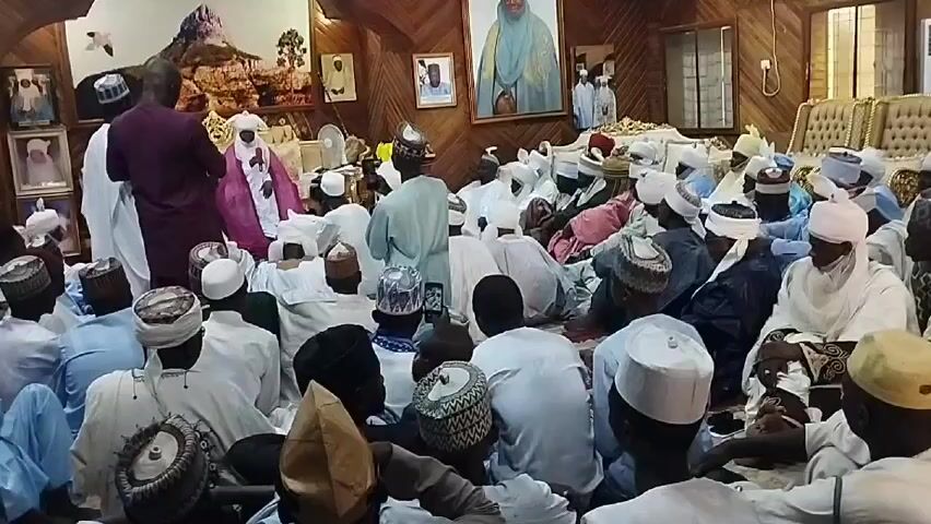 The video shows a large group of men seated on the floor in a wood-paneled room. Many of the men are wearing traditional clothing, including white robes and various types of caps. There are several framed portraits on the walls. One man in a maroon robe stands near the front of the group, while another man in a pink robe is seated among them. The room is furnished with ornate gold chairs in the background.