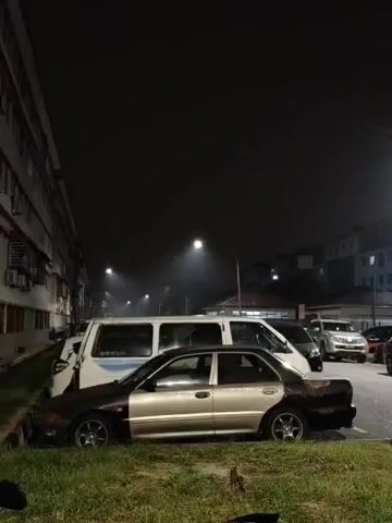 THE VIDEO SHOWS A NIGHTTIME SCENE OUTSIDE AN APARTMENT BUILDING. THERE IS A LINE OF PARKED VEHICLES, INCLUDING SEVERAL VANS AND CARS. ONE CAR IN THE FOREGROUND IS A TWO-TONED SEDAN, WITH THE FRONT HALF APPEARING TO BE A LIGHTER COLOR THAN THE REAR HALF. THE REAR HALF OF THE CAR IS DARKER AND APPEARS TO BE RUSTED OR DAMAGED. THE AREA IS LARGELY DARK, WITH STREETLIGHTS ILLUMINATING SOME AREAS AND CREATING A HAZY EFFECT. THERE IS GRASS AND SOME VEGETATION IN THE FOREGROUND. A MOTORCYCLE IS PARTIALLY VISIBLE IN THE BOTTOM LEFT CORNER OF THE FRAME. THERE ARE NO PEOPLE VISIBLE IN THE VIDEO.