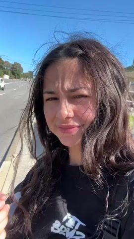 The video shows a young woman with long brown hair walking along a sidewalk next to a street with cars. She is wearing a black t-shirt and a backpack. The wind is blowing her hair around. She asks if she is crazy or if it is windy or both. The sky is blue and sunny. There are houses and trees in the background.