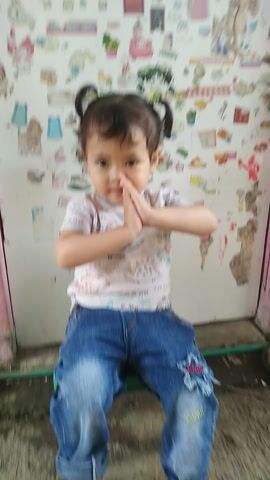 A young child with dark hair tied in two small pigtails is featured in the video. The child is wearing a white short-sleeved t-shirt with various patterns, including what appears to be dinosaurs, and blue jeans. Initially, the child has their hands pressed together in front of their chest, then they quickly smile widely and brightly at the camera, showing a cheerful and playful expression. The child is positioned low, possibly sitting or kneeling on a floor, against a light-colored wall that is extensively decorated with numerous colorful stickers. These stickers depict a variety of cartoon characters, objects, and possibly letters, giving the impression of a child's play area or room. Some of the stickers and the wall show signs of age or wear. The lighting suggests it is daytime.