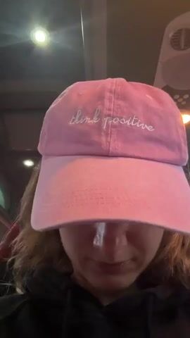 A young woman wearing a pink baseball cap with the words "think positive" embroidered on it is looking down and smiling slightly. She has on a black hoodie and is wearing white wireless earbuds. The background appears to be the interior of a vehicle, possibly a bus, with ceiling lights and a window visible. The lighting is dim.