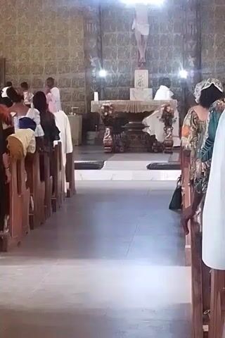 The video shows the interior of a church with people standing in pews. The focus is on the backs of the people. An altar is visible at the end of the aisle, with a figure of Jesus on the cross above it. There are flowers on the altar and candles lit on either side. Some of the people are wearing light-colored clothing, and one person is wearing a colorful, floral-patterned outfit. The church walls are a light brown color with a patterned texture. Light fixtures are visible on the ceiling. A person in the foreground is wearing a dark gray jacket.
