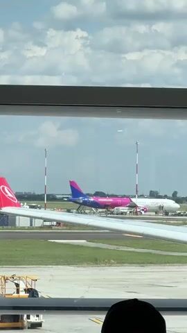 The video shows an airport tarmac viewed through a large window. In the foreground, the wing and part of the fuselage of a white airplane are visible. Behind this, two other airplanes are parked on the tarmac. One is predominantly pink with blue accents, and the other is white with some text on its side. Two tall poles with red and white stripes stand on the tarmac between the airplanes. The sky is blue with scattered white clouds. The bottom of the frame shows the heads of several people sitting, presumably passengers, facing the window. The overall scene suggests a departure or arrival gate at an airport.