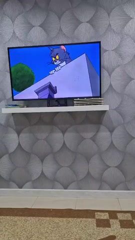 The video shows a wall with grey, circular patterned wallpaper. A flat-screen television is mounted on the wall, resting on a white shelf. The television displays a cartoon image of Tom the cat from Tom and Jerry. The image on the TV then glitches and displays distorted colors and lines, indicating a screen malfunction. Below the television, on the shelf, are several stacked items, possibly books or DVDs. The floor is covered by a carpet with a geometric pattern.