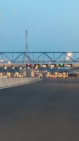 The video shows a view from a vehicle approaching a toll plaza. The sky is a pale blue, suggesting dusk or dawn. A large bridge structure is visible in the background, with a tall, slender tower extending upwards. The toll plaza has multiple lanes, each with a booth and overhead lights. Some of the lights are red, indicating closed lanes, while others are yellow or green, indicating open lanes. Cars and trucks are visible in the lanes, with some passing through the toll booths. The road surface is asphalt.