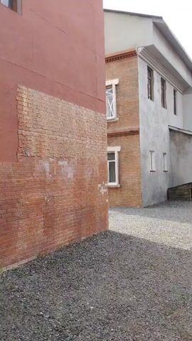 The video pans across a courtyard area between several buildings. The ground is covered in small gray gravel. On the left, a textured brick wall with a salmon-colored facade is visible. To the right, a two-story building with a brick facade and white trim on the windows is partially seen. Adjacent to this is a gray, stucco-sided building with several windows. In the background, there are some smaller, wooden structures and a red container. The sky is cloudy with patches of blue. The video appears to be filmed from a low angle, possibly handheld, and the movement is slow and deliberate.