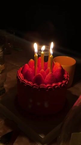 The video displays a red birthday cake, decorated with fresh strawberries and topped with four lit gold-colored candles. The cake sits on a white tray on a table in a dimly lit room. To the left of the cake, there are partially visible drinking glasses, while on the right, there is an orange mug. The scene is still and focuses on the cake, creating a celebratory and intimate atmosphere. In the background, you can hear people singing "Happy Birthday".