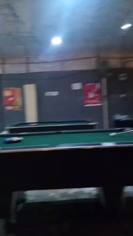 The video shows two billiard tables in a room with posters on the wall.  The room is lit by overhead lights and has a concrete floor.  One billiard table has a rack of balls set up on it. The camera moves slightly during the video.