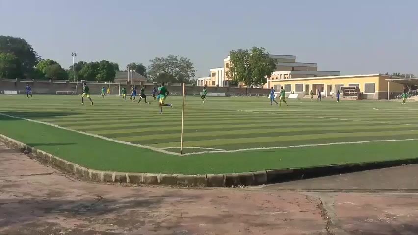 The video shows a soccer game in progress on an artificial turf field marked with white lines. Players in green and blue uniforms are actively engaged in play. A yellow corner flag stands at one corner of the field.  The field is surrounded by a low concrete barrier, with a concrete area visible between the barrier and the camera. A building with a yellow exterior can be seen in the background, along with some trees and a clear sky.