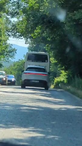 This is a first-person perspective video of a car driving on a paved road. The road is flanked by lush green trees, forming a canopy overhead. In the distance, mountains are visible under a clear blue sky, suggesting a sunny day.

The primary activity is driving. The car is following a white van and a black car. The white van appears to have "Bodyshop Travel" written on its back. The road appears to be in a rural or mountainous area, possibly a national park or scenic route, given the abundance of nature. The time of day is clearly daytime, and the weather appears to be fair. There are no people clearly visible inside the car, but the presence of multiple vehicles indicates traffic. The overall mood is serene and peaceful, fitting for a scenic drive.