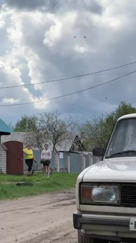 The video presents a scene in what appears to be a rural setting. The camera starts focused on two women standing near a fence, and then pans upward to capture the sky. The woman on the left wears a bright yellow shirt and dark pants, leaning against a brick gatepost. The woman on the right wears a white sweatshirt and shorts, holding a small tray or bag. In the foreground is the corner of a white car. A dirt road is visible. In the background, a wooden house with a teal-colored roof, utility poles, and trees are visible. The sky overhead is filled with dynamic, predominantly gray clouds. Numerous small flying insects are apparent throughout the video.