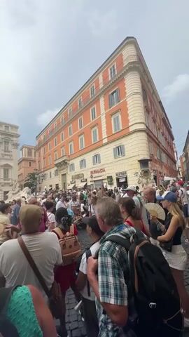 A crowded public square in front of a large ornate building. People of all ages are gathered, some looking at the building, others talking or holding up phones to take pictures. A fountain is visible to the left of the building, with water cascading down. The buildings are old, with cobblestone streets. The sky is overcast.