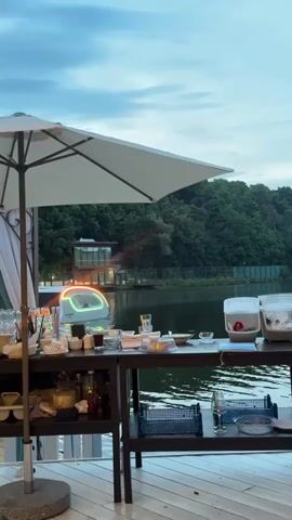 The video pans across a table set up for an outdoor event, possibly a restaurant or bar. On the table, there are various items such as glasses, bottles, food containers, and napkins. Behind the table, a body of water is visible with trees lining the opposite bank. A modern, futuristic-looking boat with illuminated accents is floating on the water. A large white umbrella is positioned over the table, providing shade. The sky is overcast, suggesting either early morning or late evening. The overall atmosphere appears serene and relaxed, with the gentle glow of the boat's lights reflecting on the water.