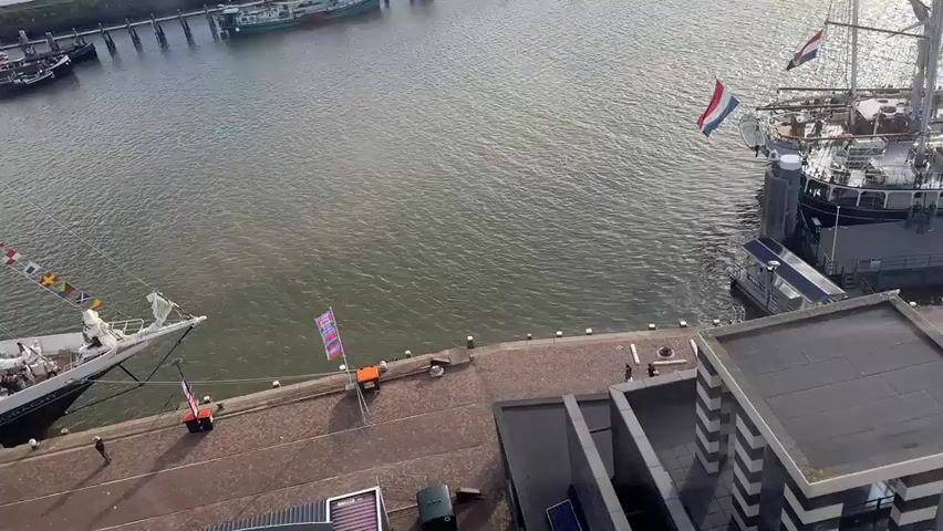 This video shows a harbor with several boats docked. A Dutch flag is visible on a ship in the background. People are walking on the pier. The water is choppy, and the sky is cloudy. The video is shot from a high vantage point, possibly a building overlooking the harbor.