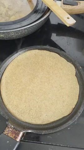 The video shows a light tan crepe cooking on a gray griddle. Steam rises from the crepe.  A plate of white crepes sits next to a small, white butter dish holding a slab of butter. A hand holding a knife cuts a pat of butter.