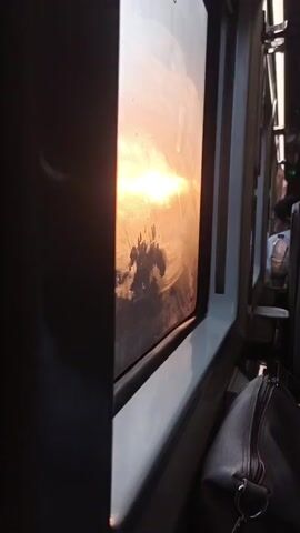 The video shows the interior of a vehicle, likely a bus or train, with a view out of a window. The sun is setting or rising, casting a warm, orange glow across the scene. The window has some condensation or smudges on it, which distort the view of the outside. In the foreground, a dark bag is resting on a seat. In the background, a few passengers are visible, seated and facing away from the camera. The overall impression is one of a quiet, contemplative journey during golden hour.