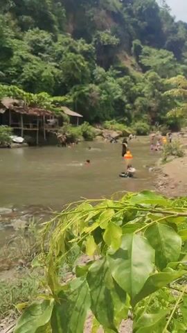 A wide shot captures a shallow river with many people swimming and playing. The riverbanks are lined with trees and vegetation, and several small wooden huts are visible on the left bank. The water is murky brown, and several people are using inflatable tubes and rings. The overall atmosphere appears to be one of leisure and recreation on a sunny day.