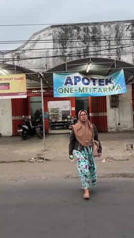 The media file shows a woman in a hijab and a floral dress walking across a street in front of a pharmacy. The pharmacy's sign reads "APOTEK ONE-MEI FARMA." The woman is carrying a white object in her right hand and a beige bag over her left shoulder. She appears to be looking towards the camera with a neutral expression.

The scene is set on a street with a paved road and a sidewalk. In the background, the pharmacy has red rolling doors and a canopy. Several motorcycles are parked in front of the pharmacy. The building behind the pharmacy has a weathered, textured facade.

The time of day appears to be late afternoon or early evening, as indicated by the diffused lighting and overcast sky. The weather seems to be cloudy. The location cues, such as the pharmacy sign and the general architecture, suggest an urban or semi-urban setting in Indonesia. The overall mood is ordinary, capturing a moment of everyday life.

Visible text includes:
- "APOTEK ONE-MEI FARMA" on the pharmacy sign.
- Text on a smaller sign on the left, partially obscured, which appears to be an advertisement, possibly for "KORONA" or similar.
- Some labels on products visible inside the pharmacy.