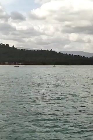 A boat is moving through the water. The water is choppy. There are clouds in the sky. There is land in the distance. There are trees on the land. There are buildings on the land. There is a pier in the water.