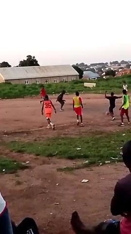 The video shows a group of young boys playing soccer on a dirt field. There are about eight boys on the field, wearing various colored shirts and shorts. The field is surrounded by grass and dirt, with a few buildings and trees in the background. There are also a few people sitting on the ground watching the game. The sky is overcast. The game appears to be a casual pickup game, with no formal markings on the field or referees. The boys are running, kicking the ball, and generally engaging in typical soccer play. The overall atmosphere seems relaxed and informal.