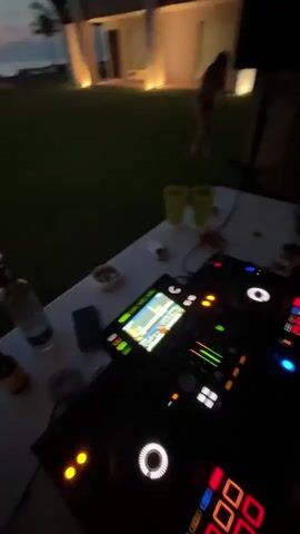 The video starts inside a room with a DJ setup featuring a digital interface with various controls and displays. It pans up to show a person, then shifts to an outdoor scene at dusk. There's a large grassy area leading to a beach with a covered gazebo-like structure with lights on inside. The sky is a mix of blue, pink, and orange, indicating either sunset or sunrise. There are some people on the beach, possibly relaxing. The video appears to be taken from a covered porch or building.