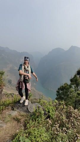 A woman stands on a rock at the edge of a mountainous landscape. She wears a green t-shirt, black leggings, white sneakers, a patterned scarf, a bandana, and a plaid shirt tied around her waist. She has a small bag slung across her body. Her arms are extended to the sides as she lifts her left leg slightly, striking a balancing pose. In the background, mountains fade into the distance, and a turquoise river winds through a valley. The sky is overcast. Vegetation covers the foreground around the rock where the woman stands.