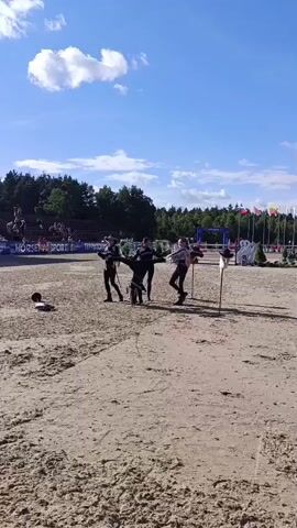 This short video captures a performance on a sandy outdoor arena, likely part of a larger equestrian event. Four individuals, dressed in black attire that suggests a historical or theatrical theme, are engaged in a coordinated act. One person, in the center, is performing a handstand with their legs extended, while the other three stand around them, holding staffs or props. They appear to be interacting with each other, possibly demonstrating a narrative or a dance routine.

The setting is a bright, sunny day with a clear blue sky dotted with fluffy white clouds. Lush green trees form a backdrop beyond the arena, and spectators can be seen seated in bleachers in the distance. Flags are visible in the background, hinting at a national or international event. The ground is covered in light brown sand, typical of a riding arena.

There is text visible on banners in the background: "SPORT" and "TERRITO...". The overall atmosphere seems festive and energetic, with the performers demonstrating skill and precision in their actions. The sunlight suggests it is midday or early afternoon. The event appears to be taking place in Ivanovskoe, Russia, as indicated by the location context.