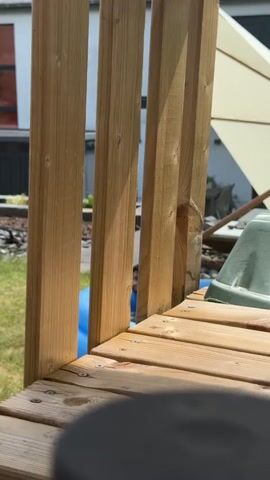 The video shows a close-up of a wooden deck. A person is partially visible behind a wooden railing, lying down in a blue object. There is a green plastic object on the deck and a tan umbrella in the background. The scene appears to be outdoors on a sunny day.