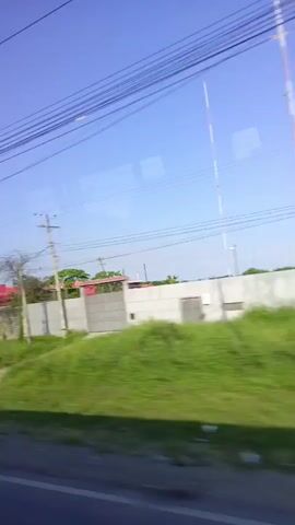 The video shows a view from a moving vehicle. The scenery includes green grass, trees, power lines, utility poles, and a long red and white building with a wall. The sky is blue. As the vehicle moves, the red building comes into clearer view. There's some debris on the ground in front of the building.