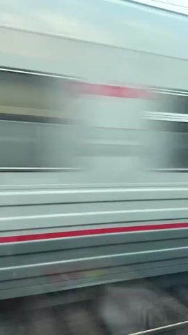 The video begins with a close-up view of a red and white train moving rapidly past the camera. The video transitions to a view from inside a moving train, showing a landscape of trees and a body of water reflecting the trees. The train tracks are visible in the foreground. As the train moves, the landscape changes to reveal some cleared land, with exposed dirt and rocks, and a small puddle of water. The scene is bright, with blue sky and scattered clouds.