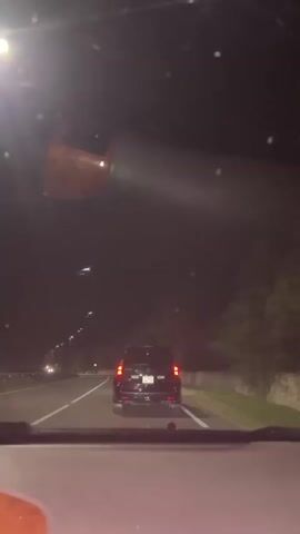 The video shows a nighttime view from inside a vehicle, looking forward through the windshield and the rearview mirror. The road ahead is illuminated by streetlights and the headlights of a black SUV in front. The SUV has its taillights illuminated and appears to be driving in the right lane of a two-lane road. Trees and foliage line the right side of the road, and a retaining wall is visible in the background. The rearview mirror reflects the interior of the vehicle, including a wooden trim element and parts of the dashboard. The overall scene depicts a car journey on a highway at night.