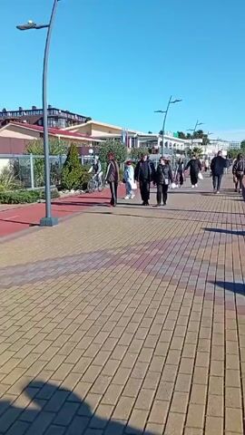 The video shows a sunny day on a seaside promenade. People are walking along a brick-paved path with a red stripe separating it from a lower level. There are lamp posts and railings along the edge of the path facing the sea. Several bicycles are parked against the railing. The sea is visible in the background under a clear blue sky. Buildings and trees can be seen on the other side of the path. People are walking in both directions. Some are standing near the railing, looking at the sea.