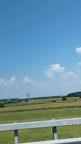 The video shows a view from a moving vehicle, likely a car or bus. The scene is a rural landscape under a clear blue sky with scattered white clouds. In the foreground, there is a silver metal guardrail. Beyond the guardrail, there are green fields, possibly agricultural land or meadows, stretching towards the horizon. In the distance, there are trees, some buildings, and tall electricity pylons with power lines. The reflections of the vehicle's interior can be seen on the windshield, particularly in the upper part of the frame, showing shapes that suggest windows or a roof. The overall impression is of a sunny day in the countryside, viewed from a moving vehicle.