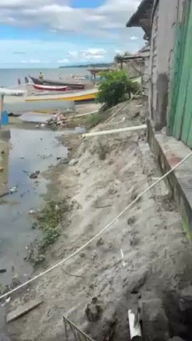 The video shows a sandy beach with several colorful boats pulled ashore. The sky is blue with puffy white clouds. People are visible in the distance, some standing on the shore and others in the water. A narrow channel of water runs across the beach, containing some debris. The camera pans slightly, revealing a building to the right, close to the water's edge.  A white pipe runs along the sandy bank near the building.  There are a few scattered objects on the beach, including what appears to be a tire, a bucket, and some construction materials. The overall scene appears to be a coastal area with signs of human activity and some litter.