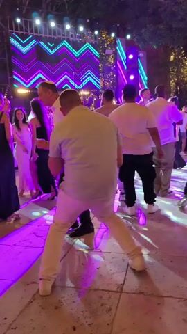 The video shows a group of people dancing at an outdoor event. There is a large LED screen in the background displaying a zig-zag pattern of colorful lights, predominantly purple and blue. The ground appears to be paved stone, and there are purple and blue lights reflecting on it. Several individuals are visible dancing. In the foreground, a man in a white shirt and black pants is dancing with his back to the camera, facing a man in a light-colored shirt and white pants. To the left, a woman in a black dress is also dancing. Further back, other people are engaged in dancing, including a woman in a white dress who appears to be the center of attention for some of the dancers. The overall atmosphere suggests a celebration or party.