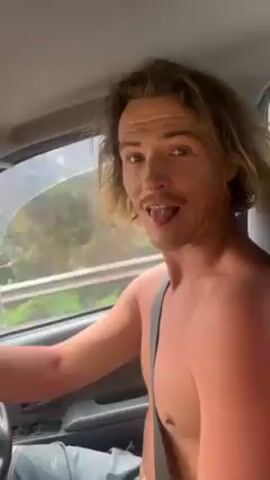 The video shows a shirtless man driving a car. He has long, light-brown hair and a mustache. He’s wearing a seatbelt and smiling, turning to face the camera and making a “shaka” hand gesture, which is typically associated with Hawaii and surf culture. He sticks out his tongue at the camera. The car is moving and the background seen through the car window shows greenery and glimpses of buildings. The steering wheel is visible.