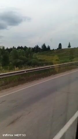 The video shows a view from a vehicle traveling along a paved road. A metal guardrail runs alongside the road, separating it from a grassy embankment. Beyond the embankment, there are trees and hills under a cloudy sky. The camera appears to be mounted inside the vehicle, as evidenced by a slight reflection on the surface. The scene is rural and depicts a typical landscape in a temperate region. The footage is brief, lasting only a few seconds, and captures a fleeting glimpse of the passing scenery. No people or specific actions are discernible in the video. The Infinix HOT 50 watermark is visible in the lower left corner of the screen, indicating the device used to record the video.