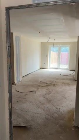 This media file captures an empty room in an unfinished state, likely undergoing construction or renovation, located in South Oxfordshire, United Kingdom. The room features bare concrete flooring with visible dust and marks. The walls are plastered and painted a light, neutral color, possibly off-white or cream, with some areas showing patches of light blue-grey primer or filler. The ceiling is also plastered.

At the far end of the room, there are white uPVC double patio doors that open to an outdoor area. Through the doors, a wooden fence and some greenery are visible, along with a glimpse of a red structure or barrier. Natural daylight enters through these doors, illuminating the room.

Several electrical elements are present: multiple power outlets are installed on the walls, and loose electrical wires hang from the ceiling and lie on the floor, indicating ongoing electrical work. There are no people visible in the image, and no discernible text. The overall impression is of a residential space awaiting further finishing touches.