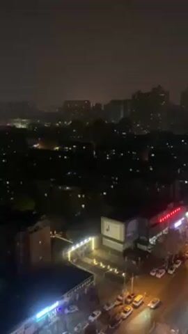 The video shows a nighttime view of a city street from a high vantage point, possibly a building. The street is illuminated by streetlights and building lights. Several parked cars line the street. Other buildings are visible in the distance, some of them high-rise apartments. The sky is dark and overcast.