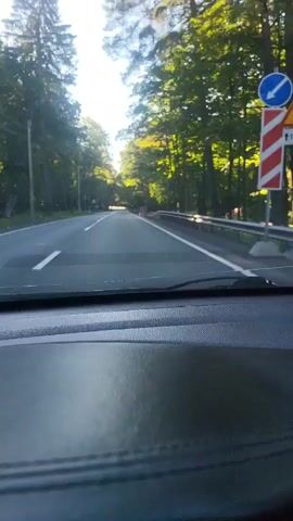 The video shows a first-person perspective from inside a car driving on a paved road. The road is lined with trees on both sides, and the sun is shining through the leaves, creating dappled light. There are white lines marking the lanes on the road. In the distance, the road curves slightly and appears to lead into a sunlit area. Traffic cones and some signage are visible along the side of the road. The dashboard of the car is visible in the foreground.